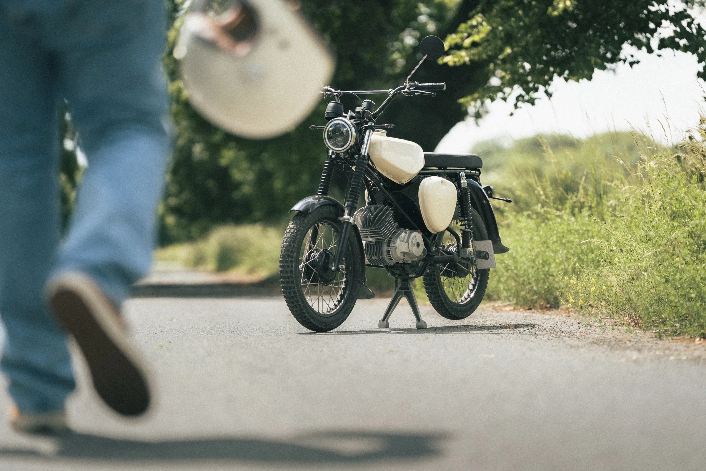 geparktes moped am straßenrand, im vordergrund unscharf eine person, die den cremefarbenen helm hochwirft