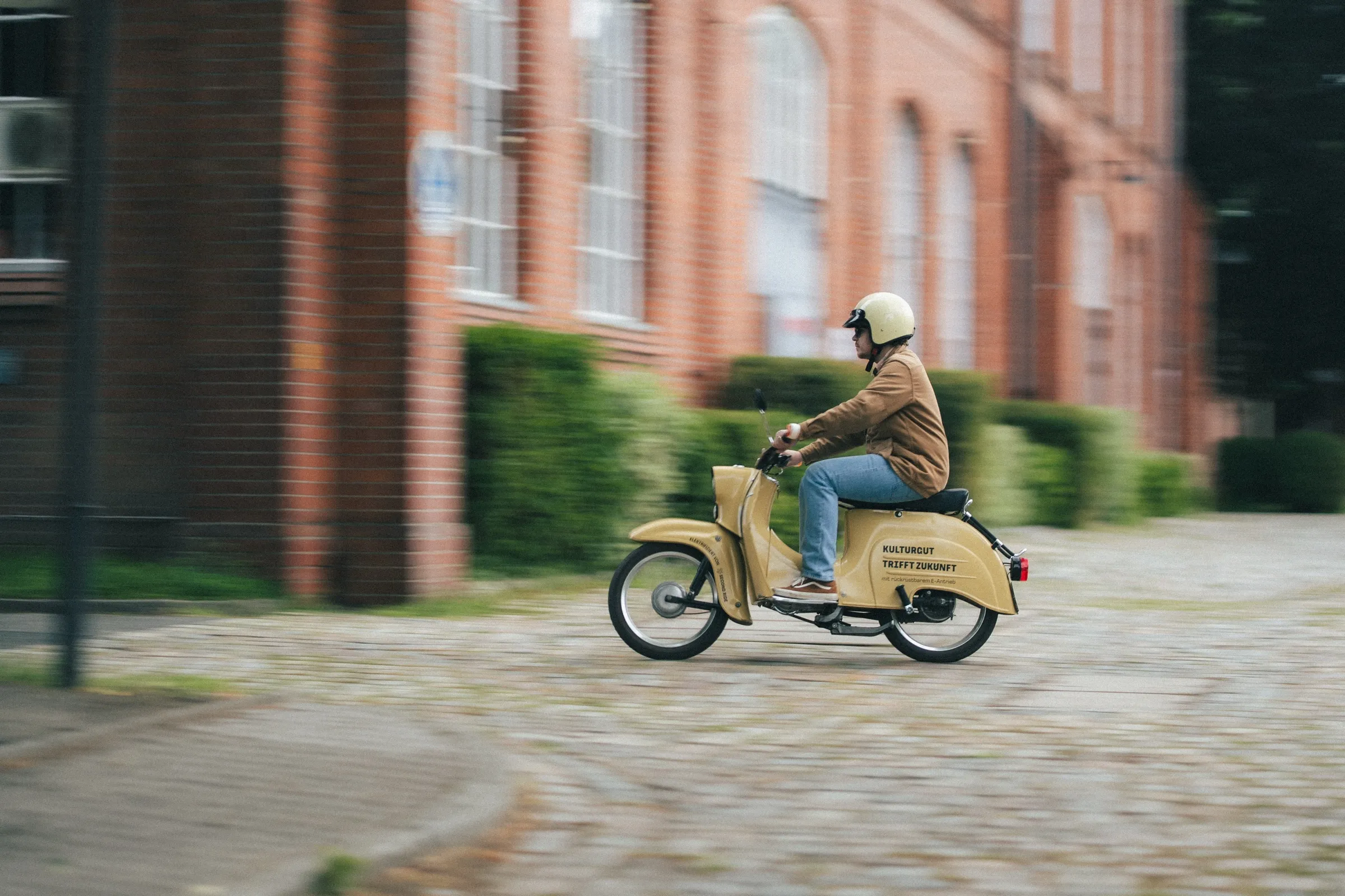 vorbeifahrender fahrer auf gelbem moped vor einer backsteinfassade in berlin