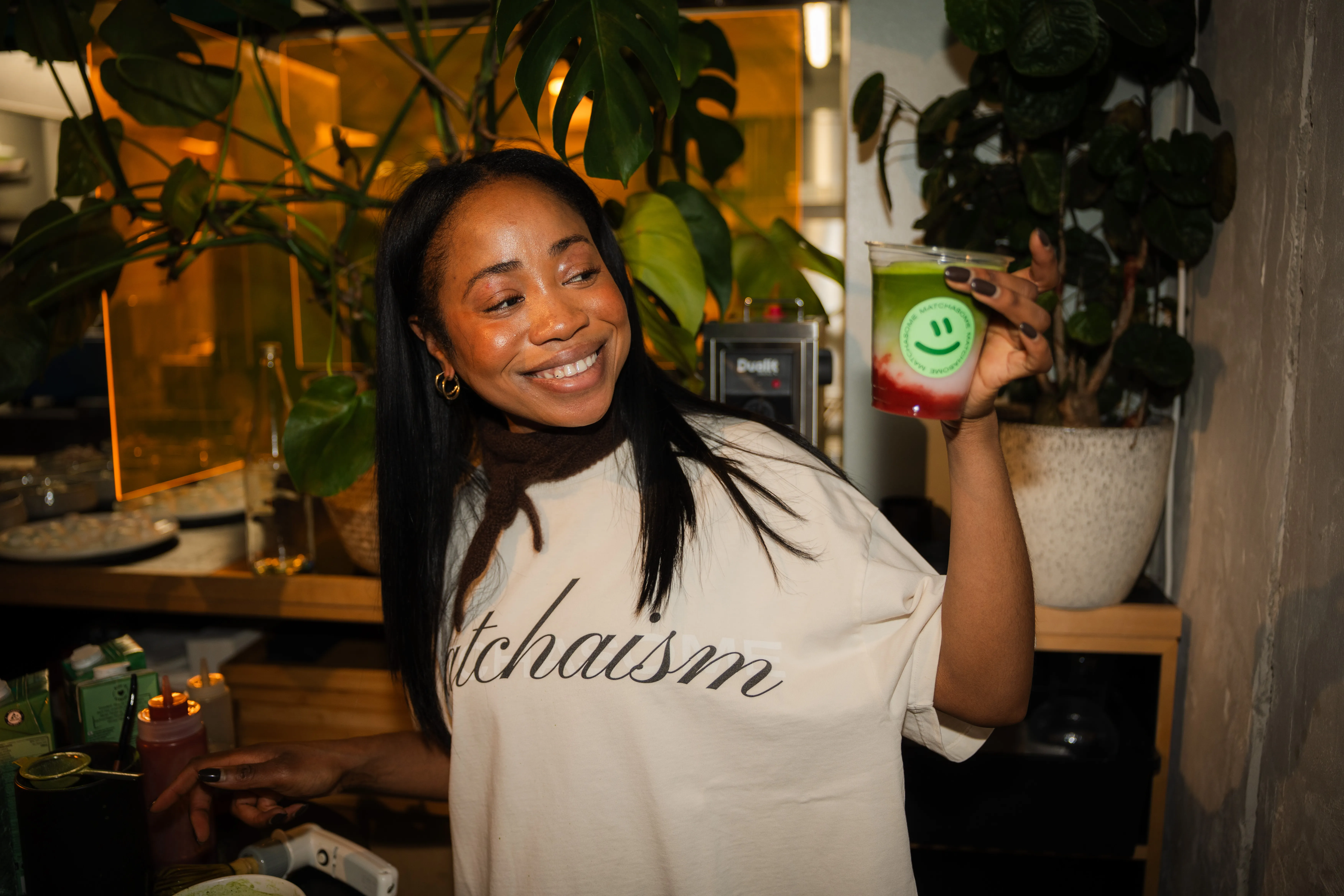 frau im „matchaisme"-shirt hält lächelnd ein grün-rotes matcha-getränk